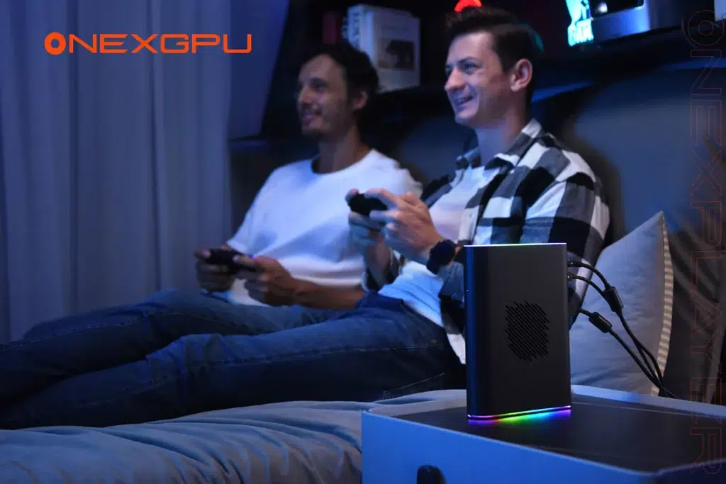 Two men sitting on a couch gaming with controllers in hand illuminated by ambient blue lighting A sleek external GPU device the ONEXGPU 2 sits on a nearby table featuring RGB lighting and multiple connected cables The ONEXGPU logo is prominently displayed