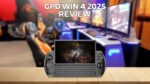 An image of the GPD WIN 4 2025 model, showcasing its compact handheld gaming device with a physical keyboard and integrated gaming controls. The device is displayed against a wooden surface in the foreground, with a blurred gaming setup featuring multiple monitors and gaming chairs in the background. The text &#039;GPD WIN 4 2025 REVIEW&#039; is prominently placed at the top of the image.