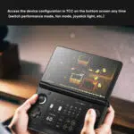 A close up first person shot of a persons hands holding an open AYN Thor handheld gaming console The top screen displays a pixel art game with a character shooting at enemies The bottom screen shows a user interface with various icons and real time performance data including a framerate of 60 FPS The text Access the device configuration in TCC on the bottom screen any time switch performance mode fan mode joystick light etc is at the top of the image