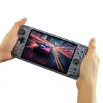 A pair of hands holding the GPD WIN 5 handheld gaming console isolated against a white background The console is dark grey and its large widescreen display shows a high speed racing game with a futuristic car speeding through a city at night with motion blur effects The ergonomic grips and all control buttons are visible