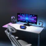 A wide shot of a modern gaming desk setup A white desk holds a large ultra wide monitor displaying a vibrant blue and purple abstract design A GPD WIN 5 handheld gaming console is propped on a stand in front of the monitor connected by a cable Other items on the desk include a futuristic speaker a headphone stand with white headphones and a chair The entire scene is lit with purple and blue LED lighting