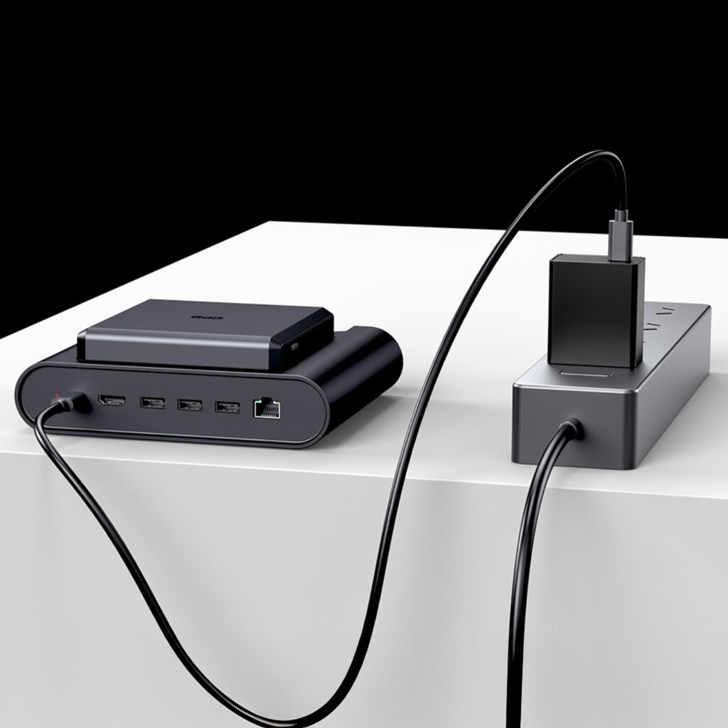 A GPD gaming dock and power hub setup on a white surface against a black and white background A dark grey rectangular dock with multiple ports is on the left with a smaller device sitting on top of it A black cable runs from the dock to a dark grey power strip or hub on the right which is also connected to a power adapter block